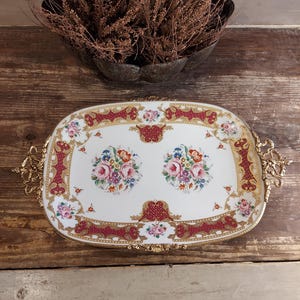 Puede incluir: Una bandeja de servir de porcelana blanca con ribetes dorados y rojos y diseños florales. La bandeja tiene dos asas doradas ornamentadas.