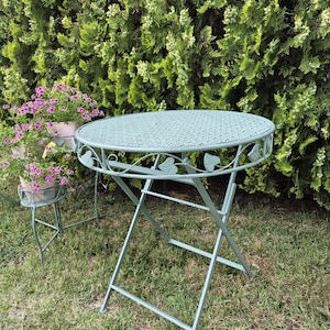 Peut inclure: Table de jardin pliante, ronde, vert clair, avec plateau texturé et motifs de feuilles. La table en métal est posée sur de l'herbe. Un petit support à plantes assorti est également visible. Diamètre : environ 60 cm.