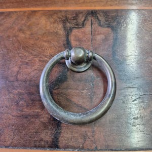 Può includere: Primo piano di una maniglia per cassetto color bronzo con maniglia circolare e base rotonda. La maniglia è montata su una superficie in legno marrone scuro con accenti di legno più chiari. L'hardware ha un aspetto vintage e antico.