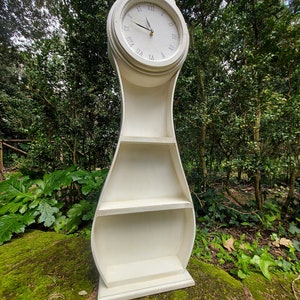 May include: A cream-coloured, freestanding clock with a decorative top and Roman numeral clock face. The clock has a curved body with two shelves. The clock is approximately 152 cm tall.