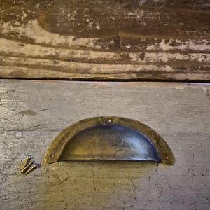Può includere: Una maniglia per cassetto a mezzaluna in stile vintage, realizzata in ottone anticato. L'hardware ha un design curvo con due fori per le viti ed è accompagnato da tre piccole viti in ottone. La maniglia è appoggiata su una superficie di legno grigio usurato.