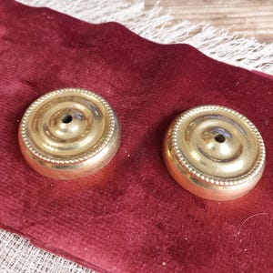 May include: Two gold-colored metal candle holders with a decorative beaded edge and a small hole in the center. The candle holders are on a red velvet surface.