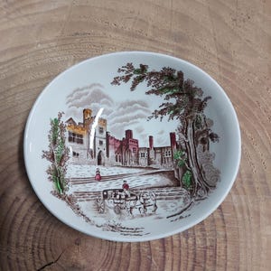 May include: A white ceramic bowl with a brown and green floral design featuring a castle and a horse-drawn carriage. The bowl has a scalloped edge.