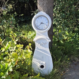 May include: A white grandfather clock with a black face and Roman numeral markings. The clock is standing in a grassy area with green foliage.