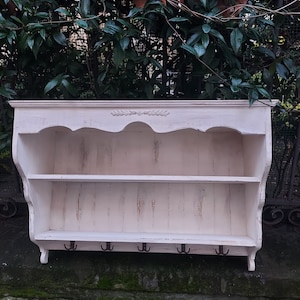 Puede incluir: Estante de pared de madera blanca con dos estantes y cuatro ganchos. El estante tiene un diseño decorativo en el borde superior y un acabado de pintura blanca desgastada.