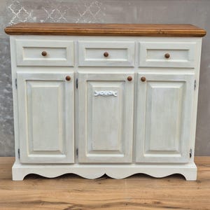 Op de afbeelding: Een witte kast met drie deuren en twee lades, met een lichtbruin houten blad en houten knoppen. De kast heeft een decoratieve, geschulpte basis en een witte, verweerde verfafwerking.