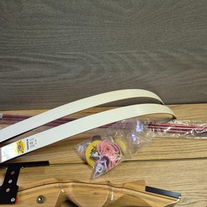 以下が含まれることがあります： 白いリムと黒い照準器を備えた木製のアーチェリー弓。弓は部分的に組み立てられており、矢や弦などの追加のコンポーネントが含まれています。弓は木製の背景に置かれています。