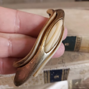 May include: A vintage, ornate, gold-toned door handle with a white, oval-shaped insert. The handle is made of metal and has a decorative, swirling design.