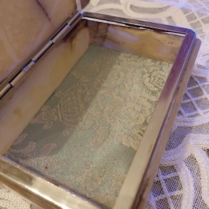 Puede incluir: Una pequeña caja rectangular con una tapa con bisagras. La caja está hecha de alabastro blanco con un borde plateado. El interior de la caja está forrado con una tela azul claro con un patrón floral.