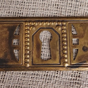 Pode incluir: Uma tampa de buraco de fechadura de latão com um design ornamentado. A tampa tem uma forma retangular com um buraco de fechadura no centro. O buraco de fechadura é rodeado por uma borda decorativa de pequenas contas. A tampa é feita de latão maciço e tem uma pátina ligeiramente envelhecida.