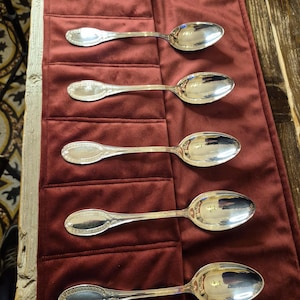 May include: Five silver spoons with ornate handles are arranged in a row on a red velvet surface. The spoons are polished and reflect light.