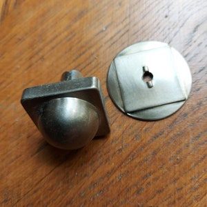 Peut inclure: Un bouton de porte de placard argenté avec un dessus rond et bombé et une base carrée. Le bouton est accompagné d'une plaque ronde assortie avec un trou carré au centre.