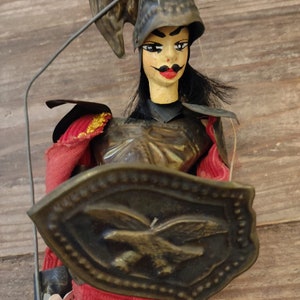 May include: A vintage puppet of a warrior wearing a helmet and armor, holding a shield with an eagle design. The puppet is made of wood and fabric and has a painted face.
