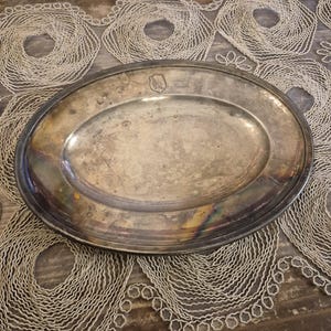 Pode incluir: Travessa oval prateada com uma borda ligeiramente elevada. A travessa tem uma aparência manchada, com um pequeno emblema perto do topo. A travessa está sobre uma toalha de renda branca sobre uma superfície de madeira.