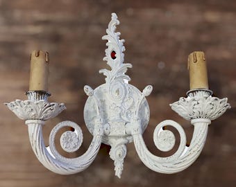Könnte beinhalten: Weiße, verzierte Wandleuchte mit zwei Armen und einer abgenutzten Oberfläche. Die Leuchte hat ein geschwungenes Design und verfügt über zwei Kerzenlampen.