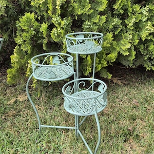 May include: A three-tiered, light green metal plant stand with decorative leaf and floral cutouts. The stand has a curved, wrought-iron style design and is set on a grassy lawn with a backdrop of green foliage.