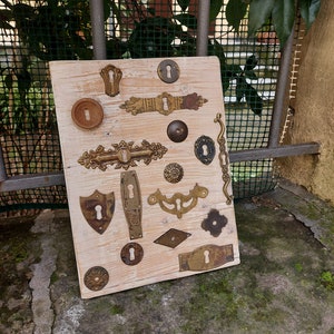 May include: A collection of antique brass keyhole escutcheons and door hardware pieces displayed on a light wood background. The pieces are arranged in a grid pattern and include a variety of shapes and sizes.
