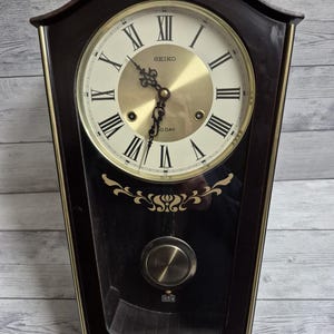 May include: A black wooden wall clock with a gold face and Roman numeral hour markers. The clock has a pendulum and says "SEIKO" and "30 DAY" on the face.