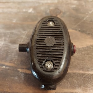 Può includere: Una spina elettrica vintage, marrone scuro, con un corpo ovale e scanalato. La spina ha un piccolo pulsante rosso su un lato e una sporgenza cilindrica sull'altro. Due viti fissano la sezione scanalata. La spina risale probabilmente alla metà del XX secolo.