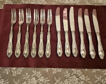 Könnte beinhalten: Ein Satz antiker Silberbestecke, darunter Gabeln und Messer, auf einem weinroten Tuch angeordnet. Das Besteck hat verzierte, detaillierte Griffe und eine polierte, reflektierende Oberfläche. Das Set wird auf einer Oberfläche mit floraler Spitze präsentiert.