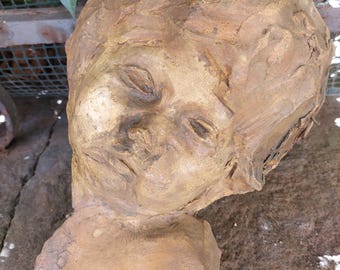 May include: A terracotta sculpture of a person's head with closed eyes. The sculpture is weathered and has a rough texture.