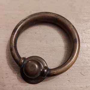 Könnte beinhalten: Ein Messing-Ringzug mit einem runden Knopf. Der Ringzug ist ein häufiges Hardware-Merkmal, das an Möbeln zu finden ist.
