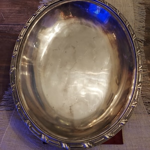 Puede incluir: Un plato de servir ovalado grande de plata con un borde decorativo elevado. El plato está hecho de metal pulido y refleja la luz.