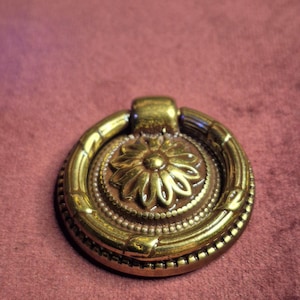Puede incluir: Un tirador de cajón de latón antiguo con un diseño floral. El tirador tiene forma circular con una flor central, rodeada de detalles de cuentas y estrías. El tirador de anillo está unido a una base rectangular.