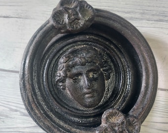 Può includere: Un batacchio per porta in ghisa nera con un design circolare che presenta un viso al centro. Il batacchio ha due maniglie decorative a forma di fiore.