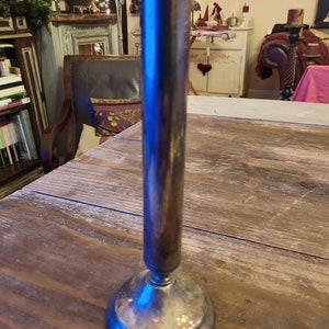 以下が含まれることがあります： 背の高い円筒形のボディと丸いベースを持つ銀のフラワーベース。ベースは磨かれた金属製で光を反射しています。