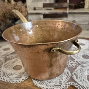 Può includere: Una pentola di rame con un manico e un manico laterale in ottone. La pentola ha un fondo arrotondato e un bordo svasato. Il rame ha un colore marrone-rossastro caldo e le maniglie in ottone hanno una tonalità dorata. La pentola è probabilmente vintage.