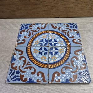 May include: Four square ceramic tiles with a repeating pattern. The tiles feature a light blue background with a central circular design in shades of blue, brown, and yellow. The design includes floral and geometric elements.
