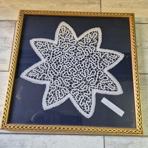May include: A framed, white, star-shaped lace doily. The doily is set against a dark blue background within a gold-colored frame. The lace has an open, intricate design. The frame is rectangular.