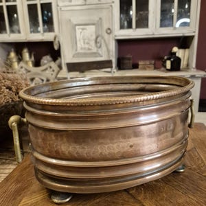 Puede incluir: Una jardinera de metal color cobre con forma rectangular redondeada. La jardinera tiene un borde decorativo, bandas horizontales y pequeños pies. Cuenta con dos asas de latón en los laterales. La jardinera se exhibe sobre una superficie de madera.