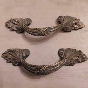 Pode incluir: Duas puxadores de gaveta de estilo antigo adornadas com um design de folha. As puxadores são de metal e têm um acabamento bronze escuro.