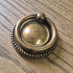 Könnte beinhalten: Ein runder, messingfarbener Schrank-Knopf mit einem perlenbesetzten Rand und einem Ring zum Ziehen.