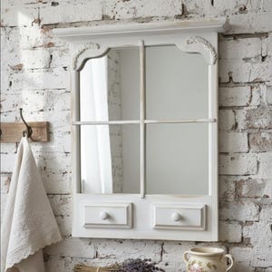 Peut inclure: Un miroir mural blanc en forme de fenêtre avec deux petits tiroirs et un dessus décoratif. Le miroir est fixé sur un mur de briques blanches. Une étagère en bois en dessous contient un bouquet de lavande et un pot en céramique floral.