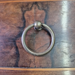 Può includere: Primo piano di una maniglia per cassetto ad anello color bronzo, con un anello circolare e una piccola base sferica. La maniglia è montata su una superficie di legno marrone scuro con venature.