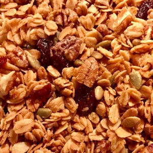 Puede incluir: Primer plano de un bol de granola con ar&aacute;ndanos, nueces de pacana y semillas de calabaza. La granola es de color marr&oacute;n dorado y los ar&aacute;ndanos son de color rojo intenso.