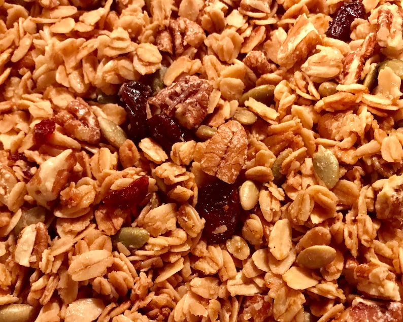 Puede incluir: Primer plano de un bol de granola con ar&aacute;ndanos, nueces de pacana y semillas de calabaza. La granola es de color marr&oacute;n dorado y los ar&aacute;ndanos son de color rojo intenso.