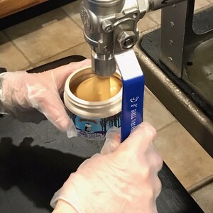 K&ouml;nnte beinhalten: Ein wei&szlig;er Plastikbeh&auml;lter mit einem blau-wei&szlig;en Etikett mit der Aufschrift "Maple Cream" wird mit einer hellbraunen Fl&uuml;ssigkeit aus einem Edelstahlventil gef&uuml;llt. Das Ventil hat einen blauen Griff mit der Aufschrift "Ball Valve".