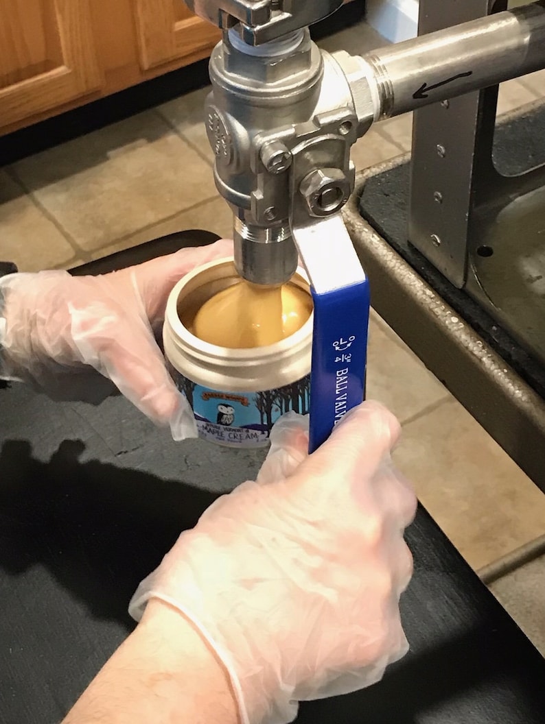 K&ouml;nnte beinhalten: Ein wei&szlig;er Plastikbeh&auml;lter mit einem blau-wei&szlig;en Etikett mit der Aufschrift "Maple Cream" wird mit einer hellbraunen Fl&uuml;ssigkeit aus einem Edelstahlventil gef&uuml;llt. Das Ventil hat einen blauen Griff mit der Aufschrift "Ball Valve".