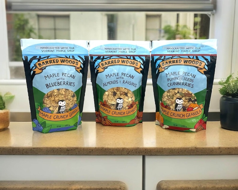 Puede incluir: Tres bolsas de granola de arce y nuez de nogal con diferentes sabores: ar&aacute;ndanos, almendras y pasas, y semillas de calabaza y ar&aacute;ndanos. Cada bolsa presenta un b&uacute;ho de dibujos animados y el texto "Barred Woods Maple Crunch Granola".