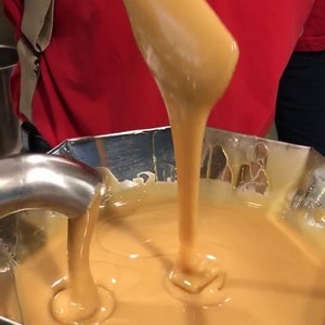 K&ouml;nnte beinhalten: Eine Person gie&szlig;t eine dicke, goldene Karamellsauce aus einem Holzl&ouml;ffel in eine Sch&uuml;ssel aus Edelstahl. Der Karamell ist glatt und gl&auml;nzend, und die Sch&uuml;ssel ist mit mehr Sauce gef&uuml;llt.