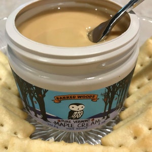 K&ouml;nnte beinhalten: Ein wei&szlig;es Glas mit Barred Woods Pure Vermont Maple Cream, eine halbe Pfund, mit einem silbernen L&ouml;ffel, der in die Creme taucht. Das Glas hat ein blaues und wei&szlig;es Etikett mit einer Eulenillustration und einer Winterwaldszene. Das Glas steht auf einem silbernen Tablett mit Crackern.