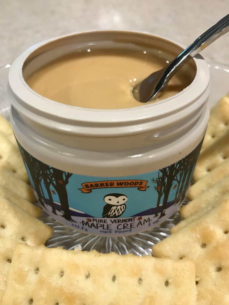 K&ouml;nnte beinhalten: Ein wei&szlig;es Glas mit Barred Woods Pure Vermont Maple Cream, eine halbe Pfund, mit einem silbernen L&ouml;ffel, der in die Creme taucht. Das Glas hat ein blaues und wei&szlig;es Etikett mit einer Eulenillustration und einer Winterwaldszene. Das Glas steht auf einem silbernen Tablett mit Crackern.