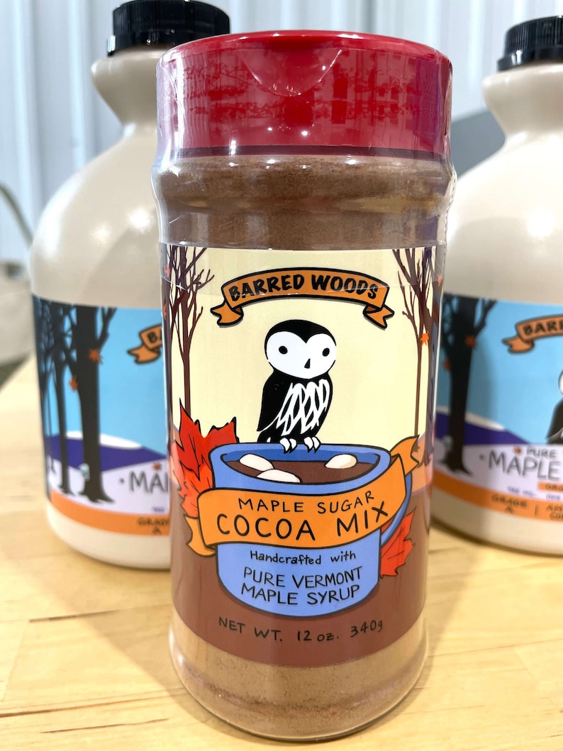 May include: A red and white jar of Barred Woods Maple Sugar Cocoa Mix. The jar features an illustration of an owl holding a mug of hot cocoa with marshmallows. The label reads "Handcrafted with Pure Vermont Maple Syrup" and "Net Wt. 12 oz. 340g".