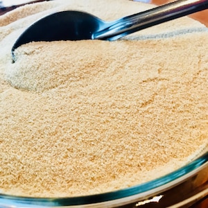 May include: A close-up of a bowl filled with a light brown, fine-grained powder. A metal spoon is partially submerged in the powder.