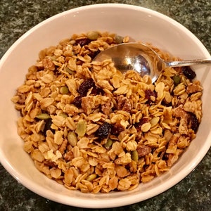 Puede incluir: Un taz&oacute;n blanco lleno de granola, con avena, nueces, ar&aacute;ndanos secos y semillas de calabaza.