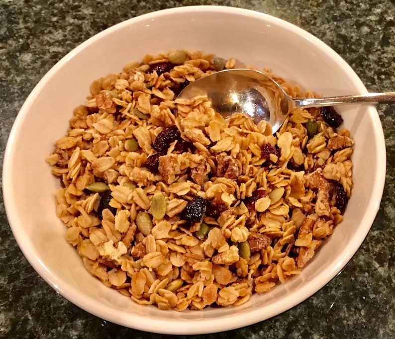 Puede incluir: Un taz&oacute;n blanco lleno de granola, con avena, nueces, ar&aacute;ndanos secos y semillas de calabaza.